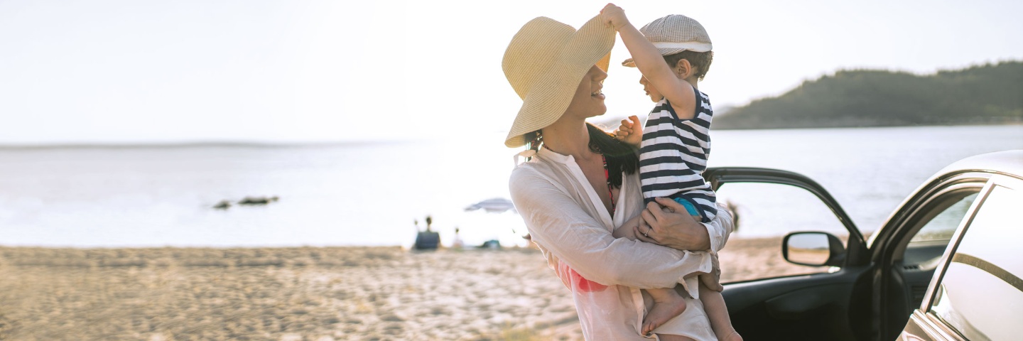 Auslandskranken-Assistance – junge Mutter mit Kind auf dem Arm am Strand. 