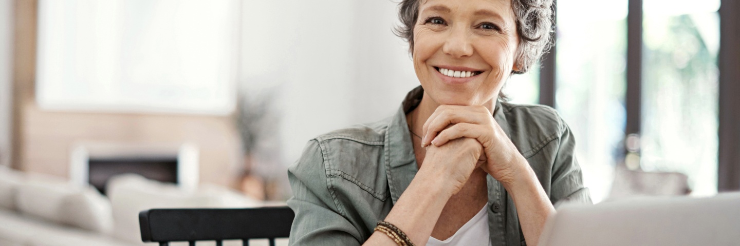 Lächelnde Frau, mit kurzen grauen Haaren, am Schreibtisch sitzend. 